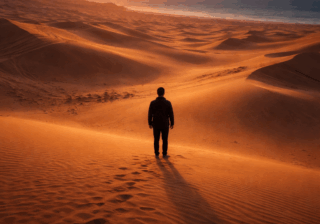 شخص وحيد يقف وسط صحراء شاسعة عند الغروب بين تلاقي الرمال والبحر في مشهد يعكس العزلة والتأمل.